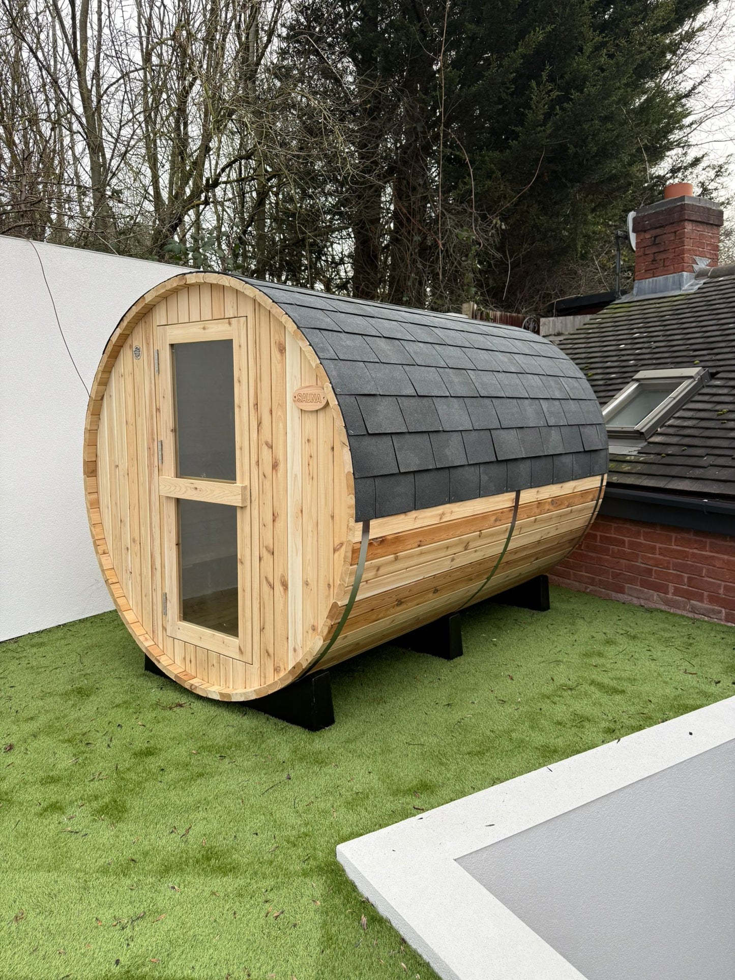 Six person barrel sauna on artificial grass beside a rendered wall